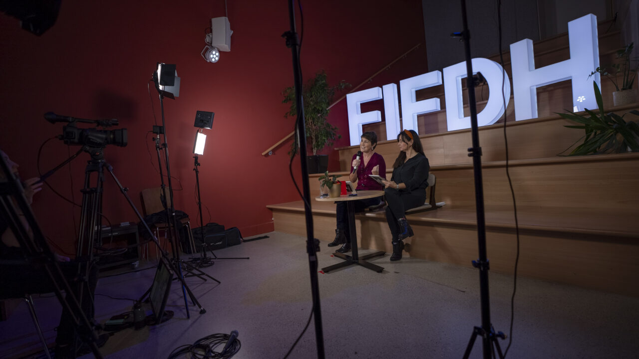 Deux femmes sont assises devant le logo FIFDH et entourées de spots lumineux. Elles parlent lors d'un webinaire en ligne.