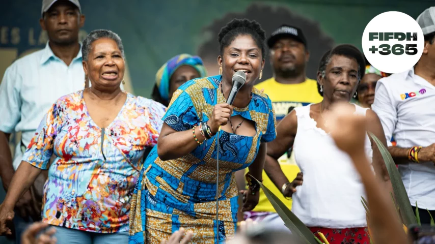 Une femme noire en tenue traditionnelle tient un micro et parle devant une foule.