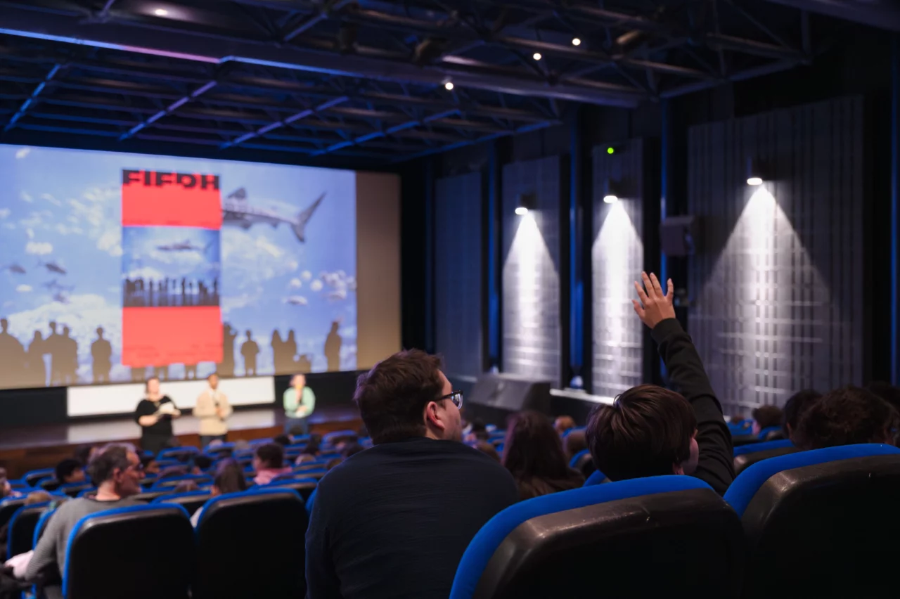 Salle de cinéma remplie, avec un élève levant la main. Le fond d'écran est l'affiche du FIFDH 2026 avec un requin